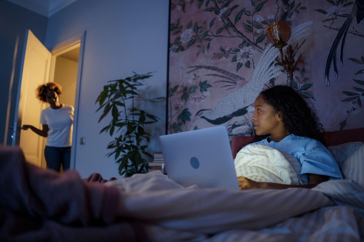a teenager staring at a glowing laptop screen in a dark bedroom reflection